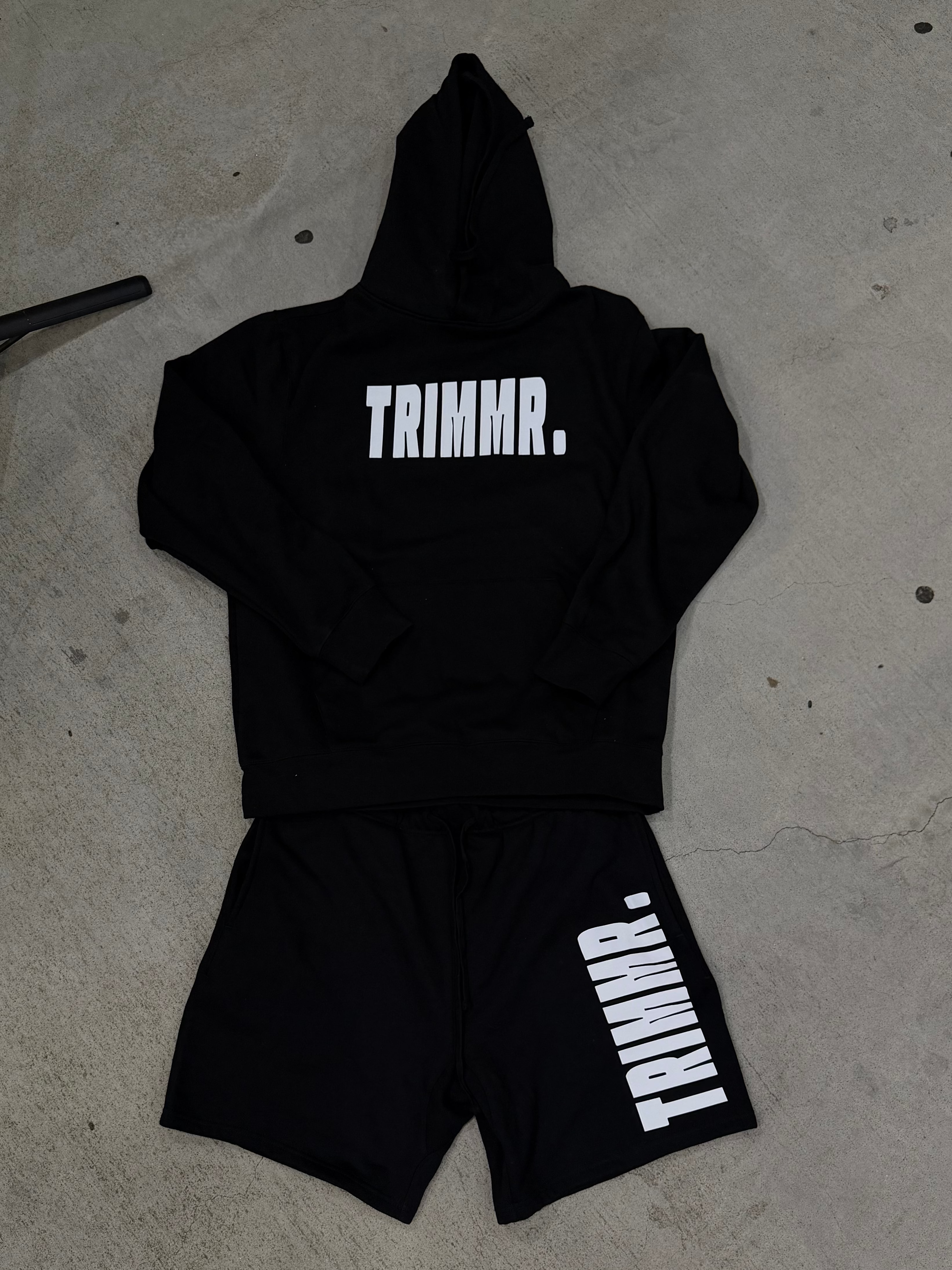 Black hoodie and shorts set with 'TRIMMR.' branding on a concrete floor.